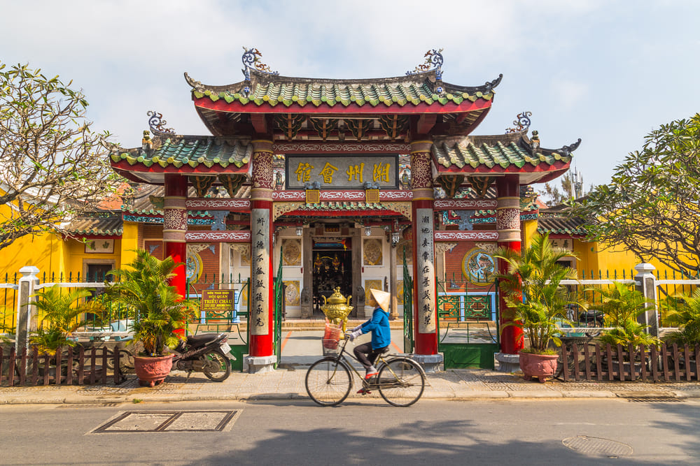 Ciclista con sombrero cónico pasando frente a un templo tradicional de fachada colorida en Hoi An, Vietnam