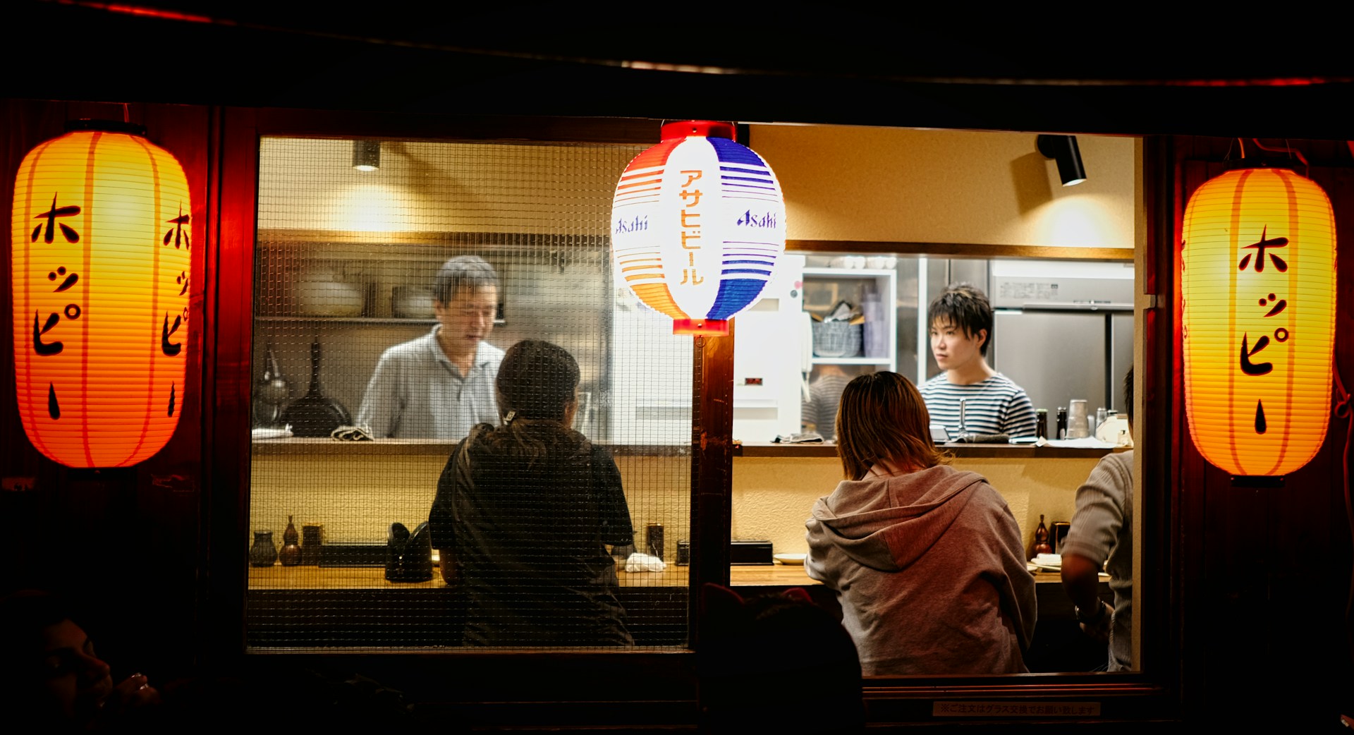 Izakaya japonés de noche visto desde la calle, con farolillos rojos y comensales en la barra